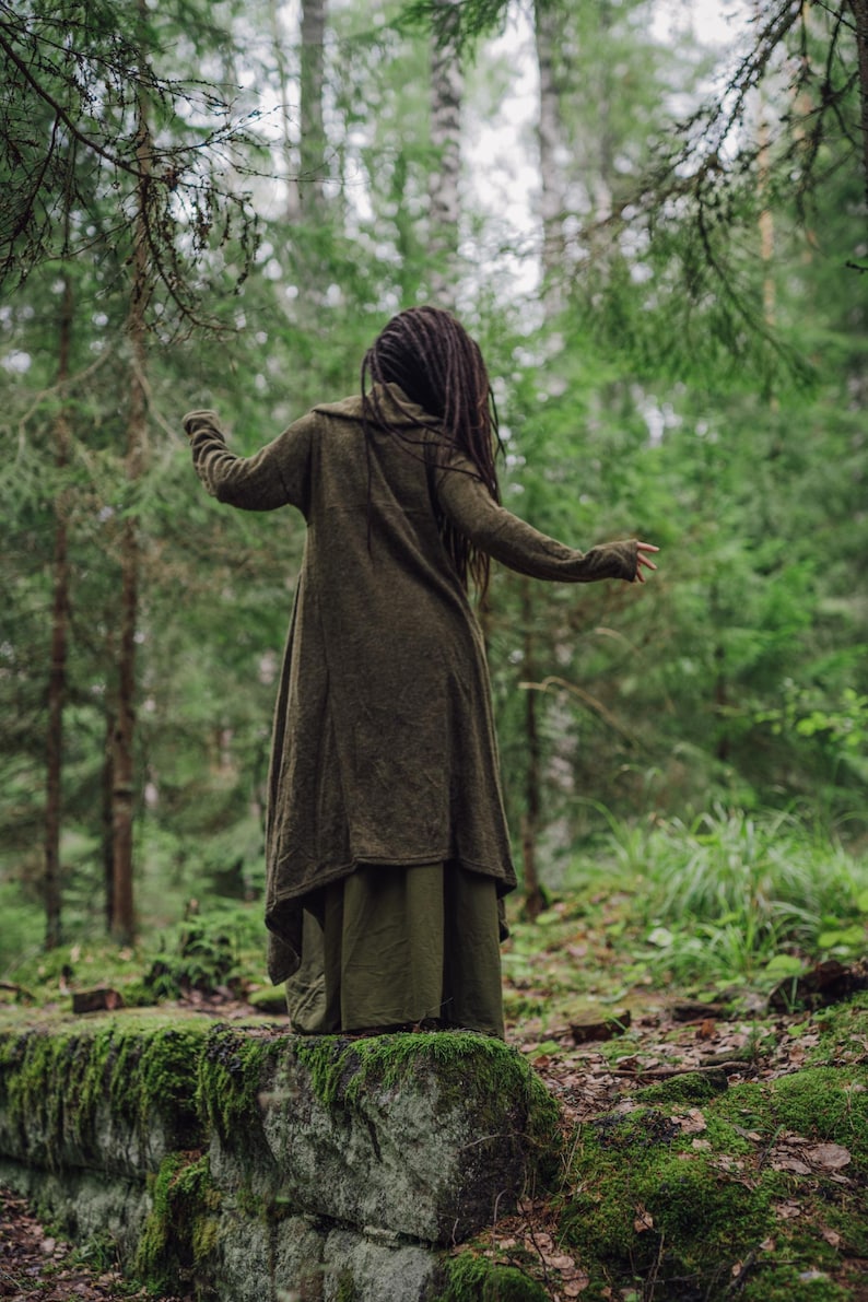 May include: A person wearing a long, dark green cardigan with a hood, standing in a forest. The person has long, dark hair and is wearing a long, dark green skirt.