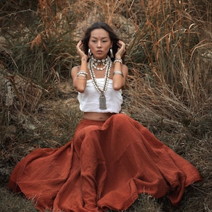 May include: A woman wearing a white top and a long, flowing rust-colored skirt, adorned with layered necklaces and bracelets. The setting is outdoors, with tall, dry grasses in the background.