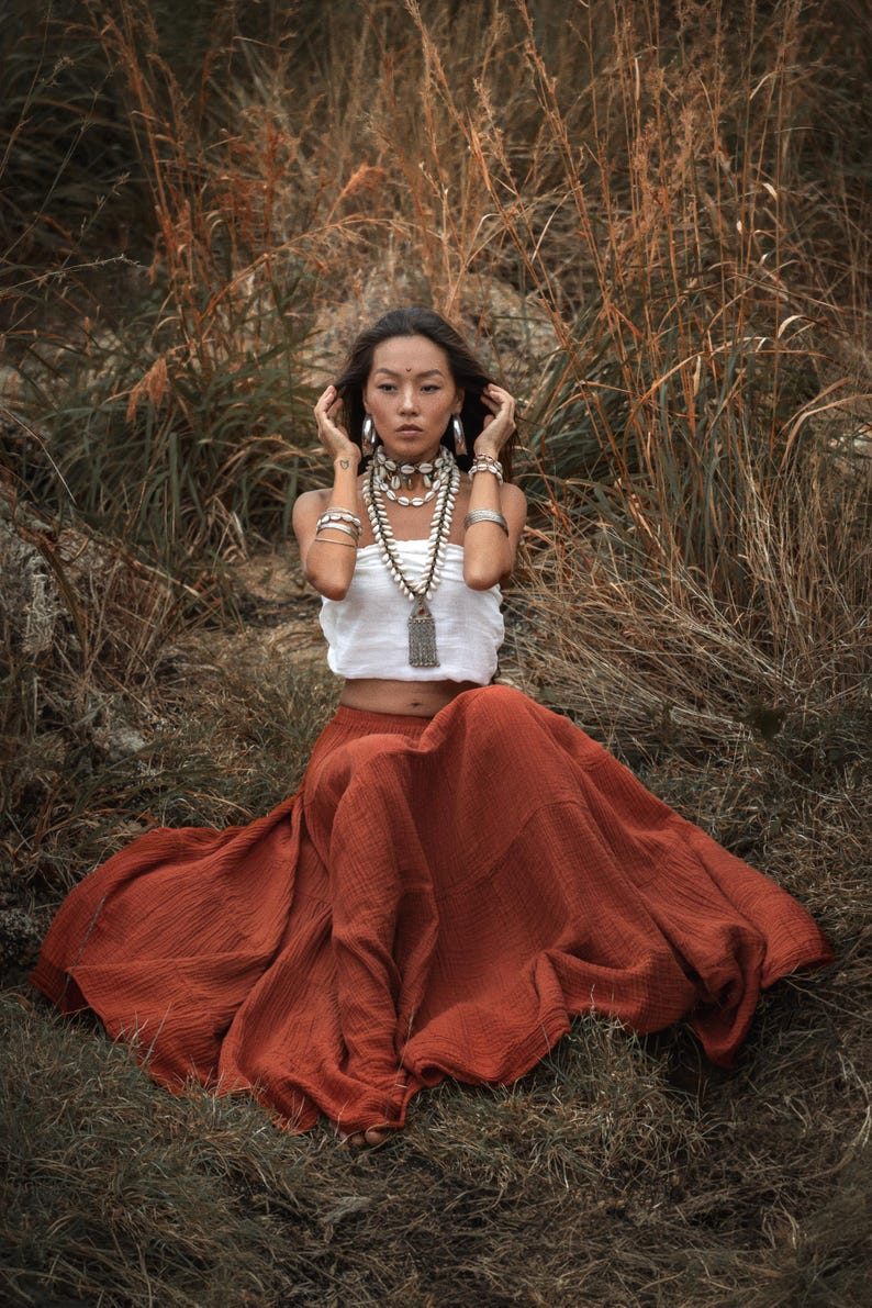 May include: A woman wearing a white top and a long, flowing rust-colored skirt, adorned with layered necklaces and bracelets. The setting is outdoors, with tall, dry grasses in the background.