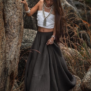 May include: A white tube top and a dark gray maxi skirt. The skirt has a tiered design. The model is wearing jewelry, including a necklace and bracelets. The model is standing near a large rock.