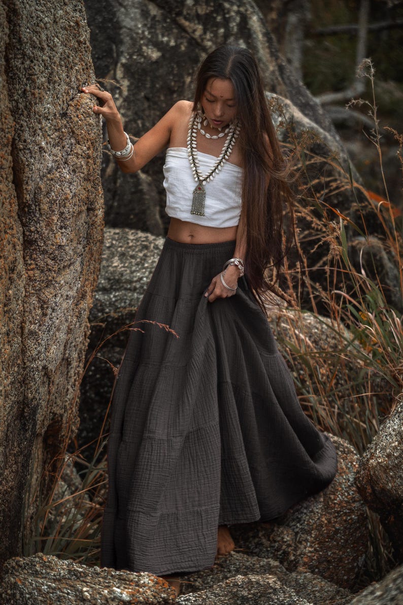 May include: A white tube top and a dark gray maxi skirt. The skirt has a tiered design. The model is wearing jewelry, including a necklace and bracelets. The model is standing near a large rock.