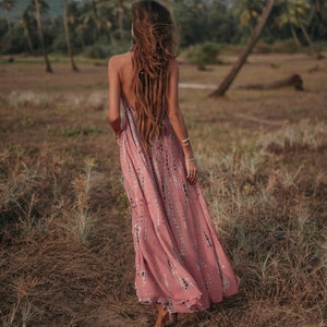 May include: A woman with long brown hair wearing a pink and white tie-dye maxi dress walks through a field of tall grass.
