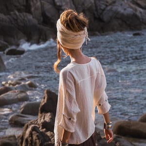 May include: A person wearing a cream-coloured, long-sleeved top with buttoned cuffs and a brown skirt, standing near a rocky shoreline. They have a head wrap and silver bracelets. The background features a rocky cliff and ocean.