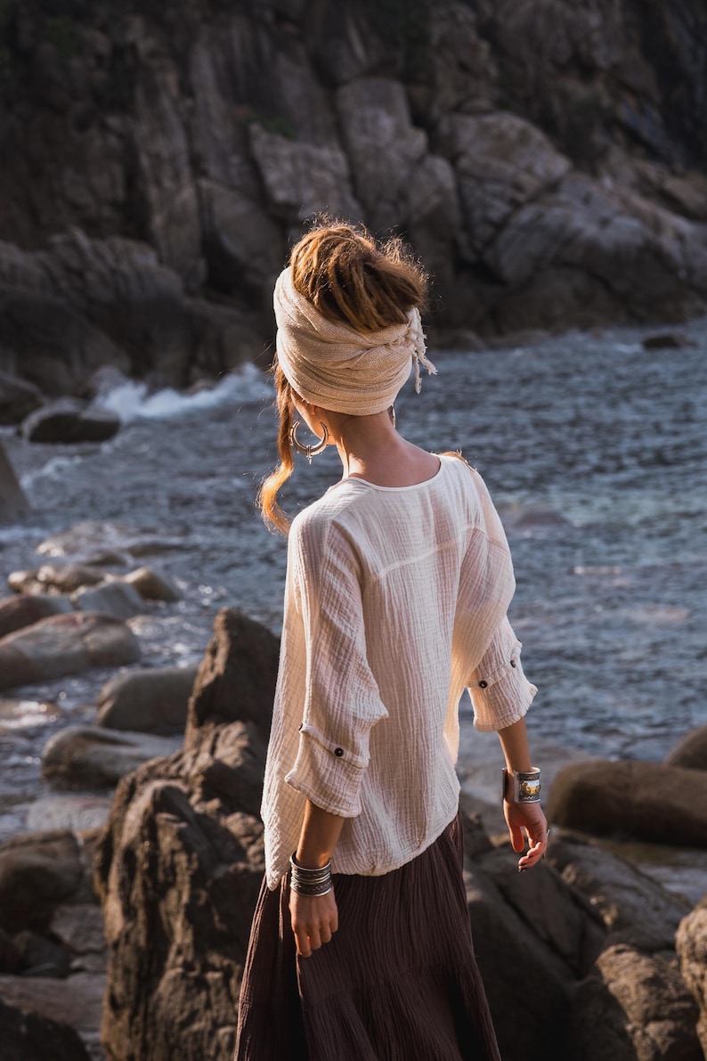 May include: A person wearing a cream-coloured, long-sleeved top with buttoned cuffs and a brown skirt, standing near a rocky shoreline. They have a head wrap and silver bracelets. The background features a rocky cliff and ocean.