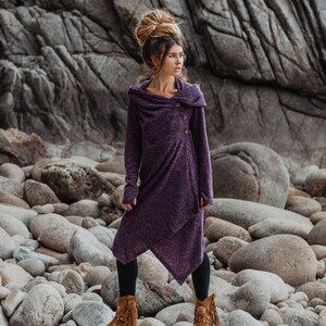 Puede incluir: Una mujer con un vestido de su&eacute;ter con capucha morado con un solo cierre de bot&oacute;n, de pie en una playa rocosa. Lleva botas marrones con detalles de flecos.