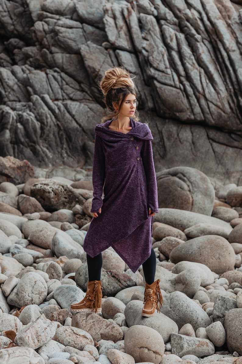Puede incluir: Una mujer con un vestido de su&eacute;ter con capucha morado con un solo cierre de bot&oacute;n, de pie en una playa rocosa. Lleva botas marrones con detalles de flecos.
