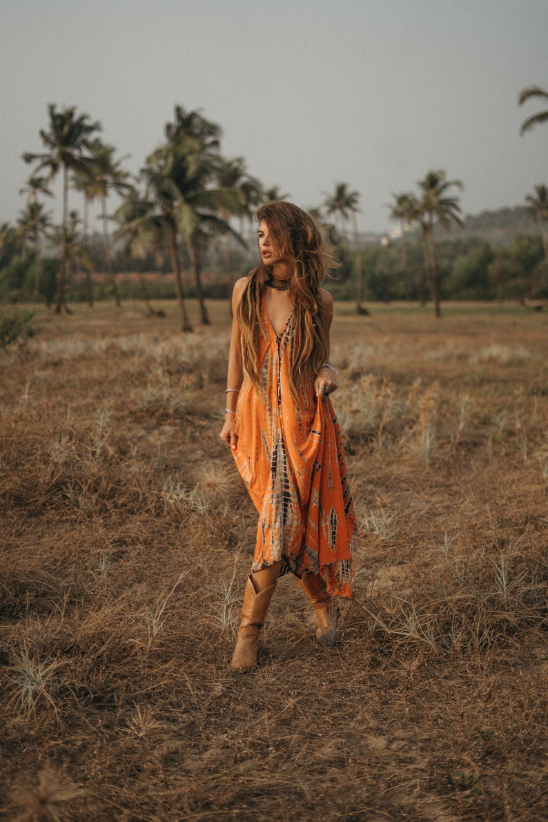 May include: A woman wearing a long orange and black tie-dye dress and brown cowboy boots walks through a field of dry grass.