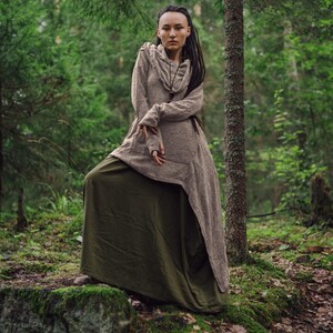 May include: A woman with dreadlocks wears a long, brown hooded tunic over a green skirt. She stands in a forest setting with green trees and moss-covered rocks.