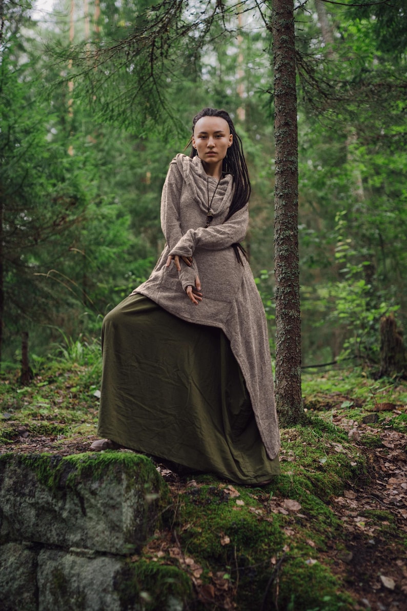 May include: A woman with dreadlocks wears a long, brown hooded tunic over a green skirt. She stands in a forest setting with green trees and moss-covered rocks.