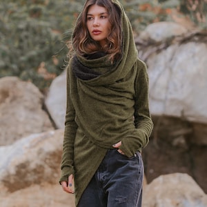 Puede incluir: Jersey con capucha verde oliva con mangas largas y cuello alto en capas. El suéter tiene un frente cruzado. El modelo lleva vaqueros negros desgastados. El fondo presenta rocas y follaje.