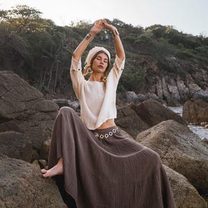 May include: A woman wearing a cream-colored top and a long, brown skirt sits on rocks near the shore. She has a matching head wrap and silver bracelets. Her arms are raised above her head.