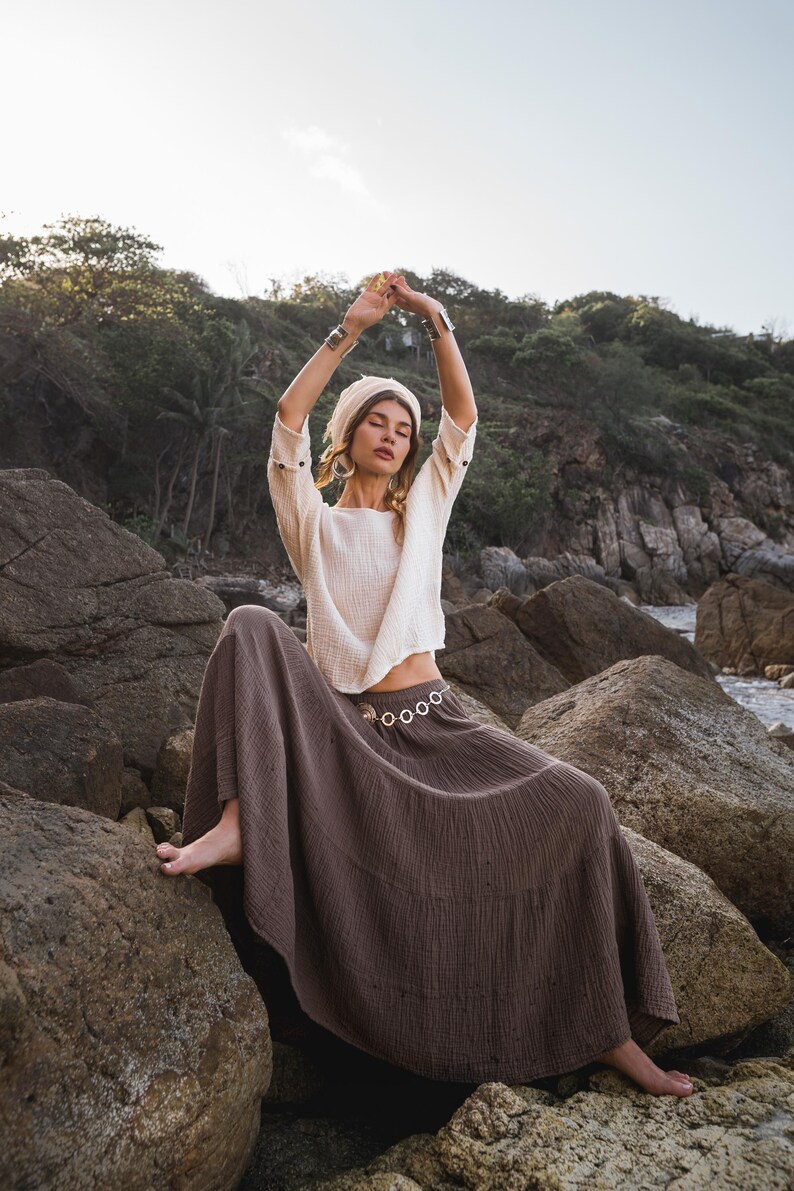 May include: A woman wearing a cream-colored top and a long, brown skirt sits on rocks near the shore. She has a matching head wrap and silver bracelets. Her arms are raised above her head.