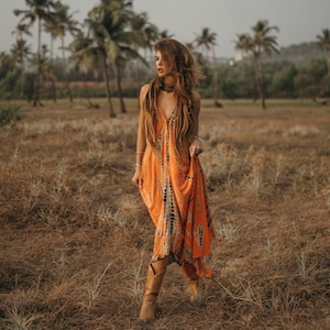 May include: A woman wearing a long orange and black tie-dye dress and brown cowboy boots walks through a field of dry grass.