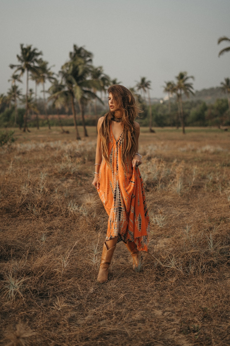 May include: A woman wearing a long orange and black tie-dye dress and brown cowboy boots walks through a field of dry grass.