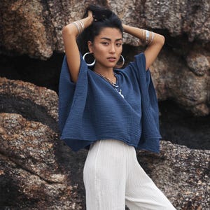 Puede incluir: Una mujer con una blusa azul oscuro holgada y un pantalón blanco de piernas anchas. Lleva pendientes de aro plateados y pulseras.