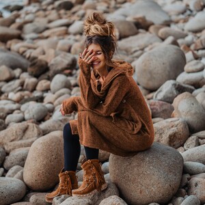 Puede incluir: Una mujer con un vestido de su&eacute;ter marr&oacute;n y botas de gamuza marr&oacute;n se sienta en una roca grande rodeada de rocas m&aacute;s peque&ntilde;as. Ella sonr&iacute;e y tiene la mano cubri&eacute;ndose la cara.