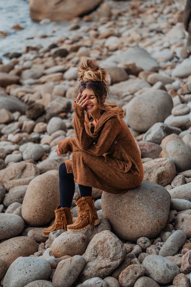 Puede incluir: Una mujer con un vestido de su&eacute;ter marr&oacute;n y botas de gamuza marr&oacute;n se sienta en una roca grande rodeada de rocas m&aacute;s peque&ntilde;as. Ella sonr&iacute;e y tiene la mano cubri&eacute;ndose la cara.