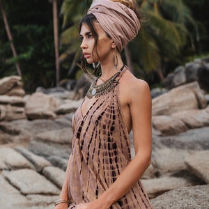 May include: A woman wearing a brown and white patterned dress with a brown head scarf and silver jewelry. She is sitting on a rock with a tropical background.