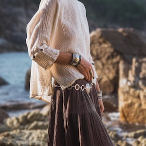 May include: A cream-coloured, long-sleeved top with buttoned cuffs and a brown tiered skirt. The outfit is accessorised with silver bracelets and a belt. The clothing has a flowing, bohemian style, suitable for casual wear.