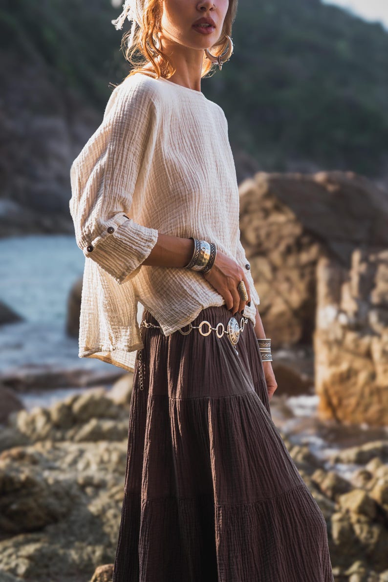 May include: A cream-coloured, long-sleeved top with buttoned cuffs and a brown tiered skirt. The outfit is accessorised with silver bracelets and a belt. The clothing has a flowing, bohemian style, suitable for casual wear.