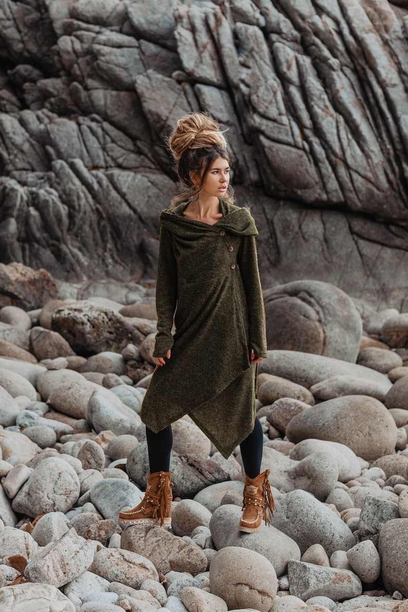 Puede incluir: Una mujer con un vestido largo de punto verde con capucha y un solo bot&oacute;n de cierre. Est&aacute; de pie en una playa rocosa y lleva botas marrones con flecos.