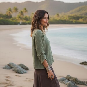 May include: A woman on a sandy beach wears a sage green top and a long brown skirt. The top has three-quarter sleeves. She is wearing silver bracelets and a ring. The background features the ocean, mountains, and palm trees.