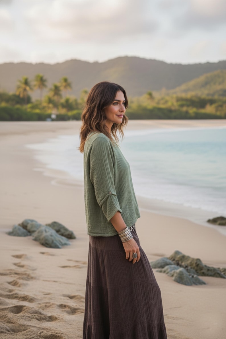 May include: A woman on a sandy beach wears a sage green top and a long brown skirt. The top has three-quarter sleeves. She is wearing silver bracelets and a ring. The background features the ocean, mountains, and palm trees.