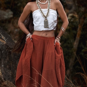 May include: A woman wearing a white tube top and a tiered rust-colored skirt. She is adorned with shell necklaces and silver bracelets. The long, flowing skirt adds a bohemian flair to the outfit, perfect for a summer look.