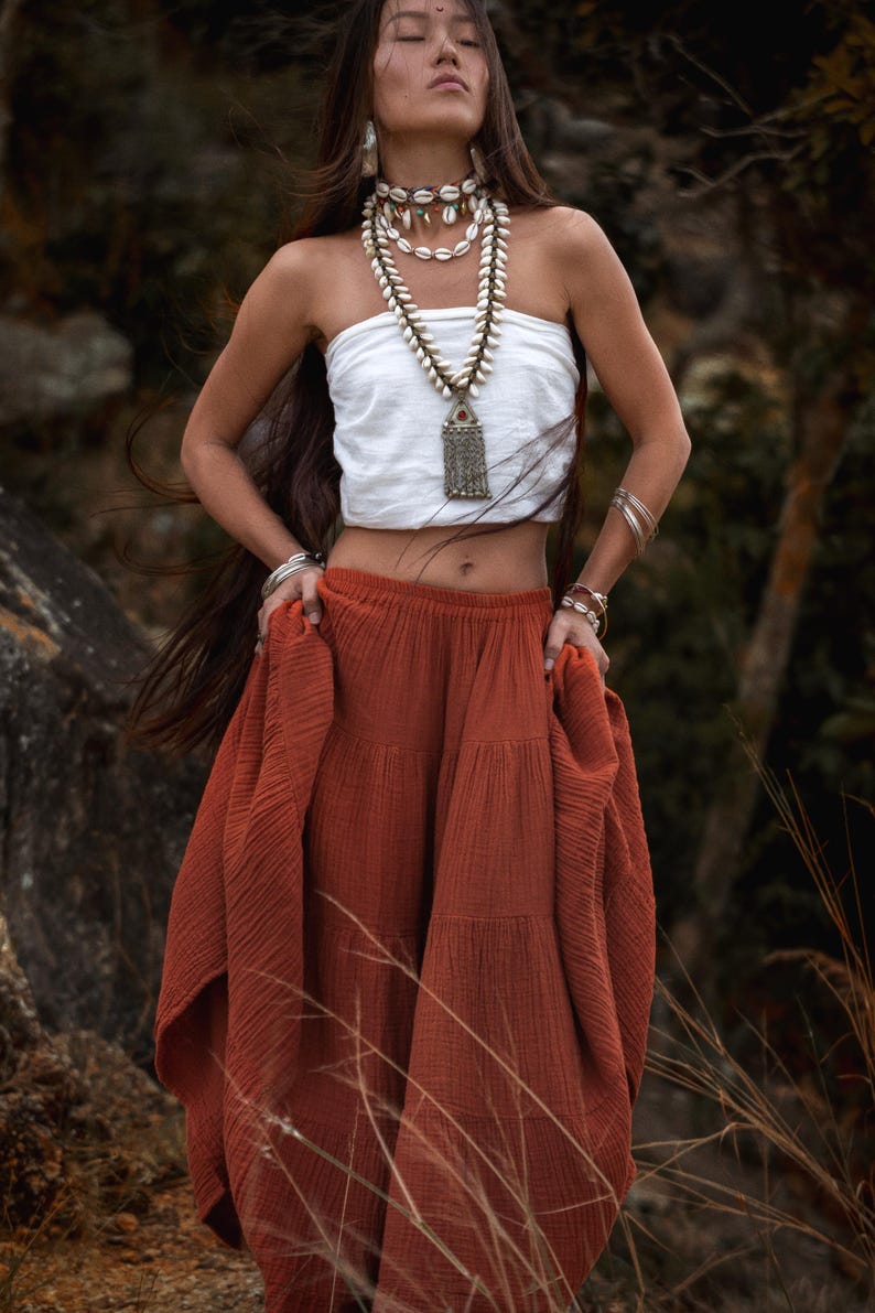 May include: A woman wearing a white tube top and a tiered rust-colored skirt. She is adorned with shell necklaces and silver bracelets. The long, flowing skirt adds a bohemian flair to the outfit, perfect for a summer look.