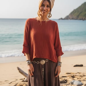 May include: A woman in a rust-coloured long-sleeved top and a brown maxi skirt. She wears a beige head wrap, large hoop earrings, and silver bracelets. A decorative belt with a pendant hangs from her waist. The background is a beach with the ocean.