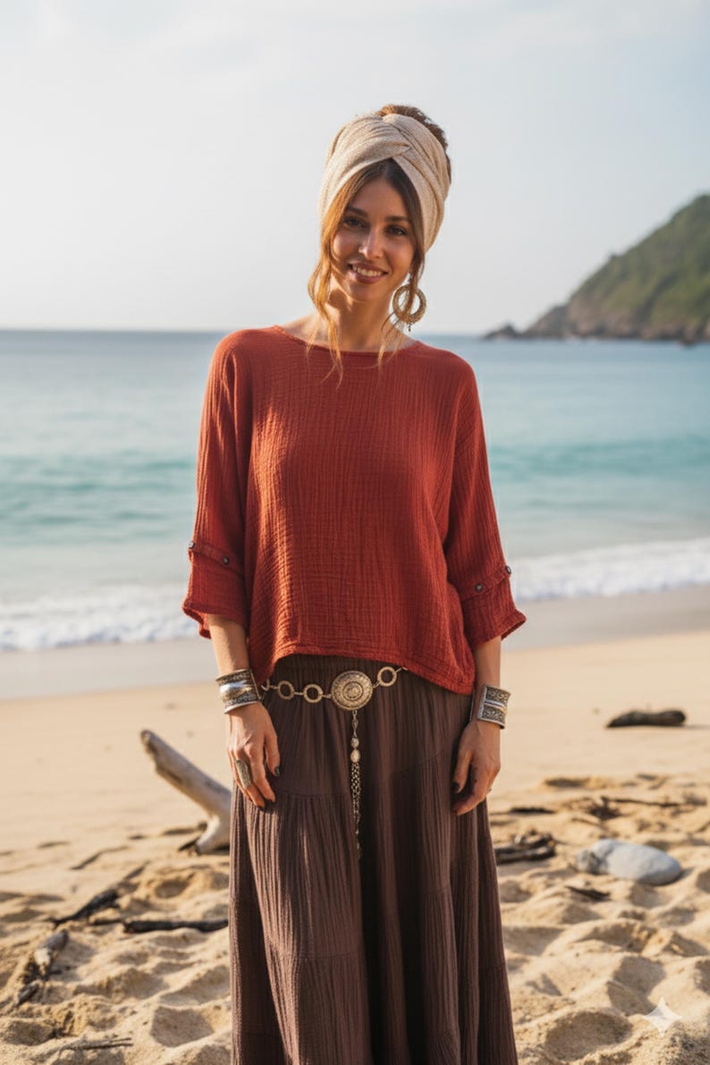 May include: A woman in a rust-coloured long-sleeved top and a brown maxi skirt. She wears a beige head wrap, large hoop earrings, and silver bracelets. A decorative belt with a pendant hangs from her waist. The background is a beach with the ocean.