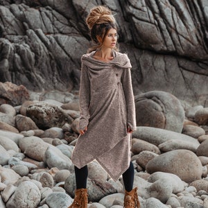Puede incluir: Una mujer con un su&eacute;ter largo de color marr&oacute;n con capucha y un solo bot&oacute;n de cierre. Est&aacute; de pie en una playa rocosa y lleva botas marrones con flecos.