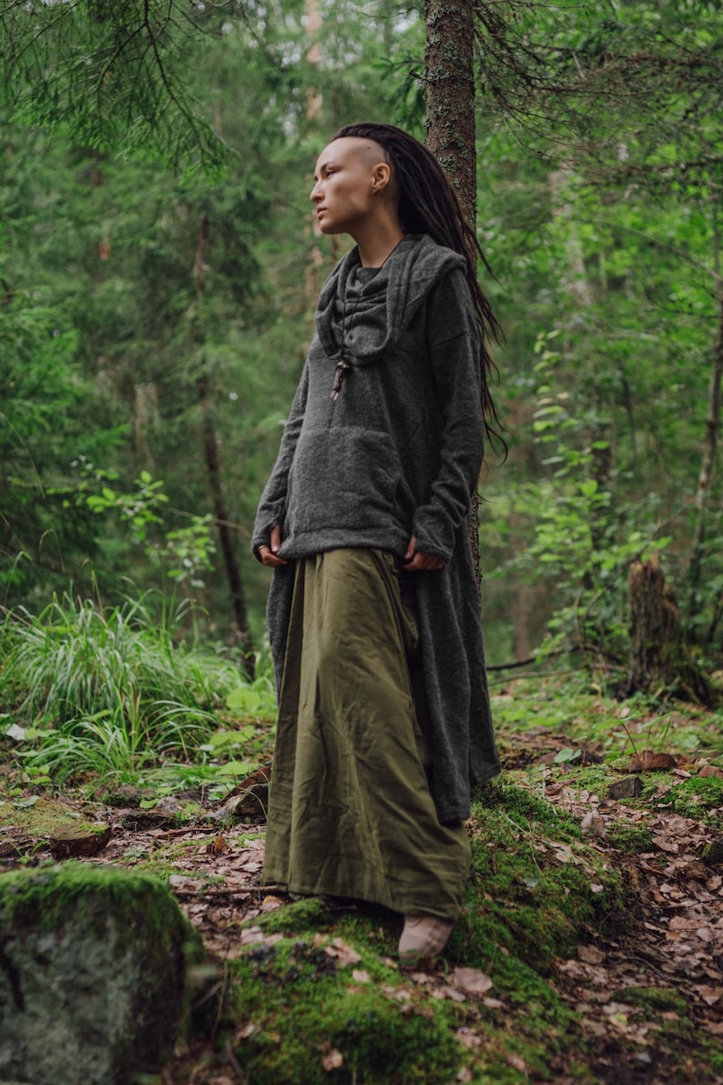 May include: A person with dreadlocks wearing a gray hooded sweater and a long green skirt standing in a forest.