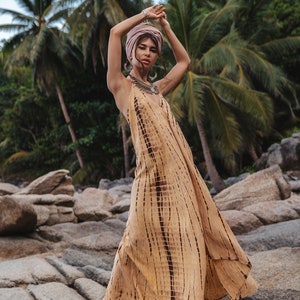 May include: A woman wearing a long, flowing, yellow and brown tie-dye dress with a pink headscarf and silver jewelry. She is standing on a rocky beach with palm trees in the background.