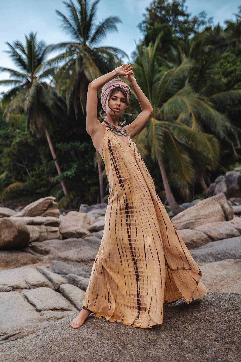 May include: A woman wearing a long, flowing, yellow and brown tie-dye dress with a pink headscarf and silver jewelry. She is standing on a rocky beach with palm trees in the background.