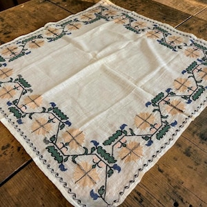 May include: A square, off-white linen tablecloth with embroidered floral and leaf patterns in peach, green, and blue. The border features a scalloped edge, adding a touch of elegance to the design. The tablecloth is placed on a wooden surface.