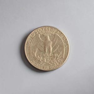 Puede incluir: Una moneda de veinticinco centavos de dólar estadounidense, con un águila con las alas extendidas y las palabras "United States of America" y "Quarter Dollar". La moneda es de color dorado claro.
