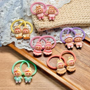 May include: A collection of colorful hair ties featuring small, cartoonish figures in various outfits. The hair ties are in shades of pink, yellow, green, and orange, with the figures in matching colors. The background includes lace and a wooden tray.