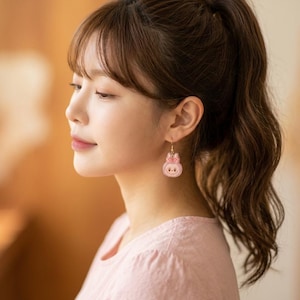 May include: A woman wearing pink bunny-shaped earrings with a bow. The earrings are hanging from gold-colored hooks. She is wearing a pink shirt with a floral design. Her hair is pulled back in a ponytail.