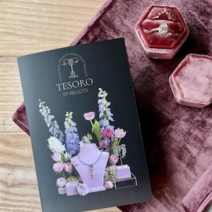 Könnte beinhalten: Eine schwarze Broschüre mit dem Text "TESORO DI VELLUTO" und floralen Illustrationen, neben einer Samt-Ringbox und einer Samtoberfläche. Ein Ring mit einem birnenförmigen Diamanten wird in der Ringbox ausgestellt.