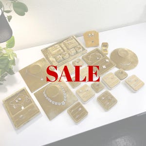 May include: An assortment of jewelry display cases and stands in various shapes and sizes, showcasing necklaces, earrings, rings, and bracelets. The displays are a muted gold color, and the word "SALE" is prominently displayed in red.