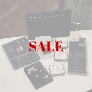 May include: Various pieces of jewellery, including necklaces, earrings, and rings, are displayed on black velvet trays and stands. The word "SALE" is in red across the image. The jewellery is presented in white and grey boxes.