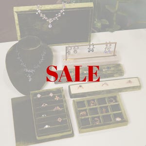 May include: An assortment of jewelry, including necklaces, earrings, and rings, displayed on green velvet stands and trays. The jewelry features various designs and gemstones. The word "SALE" is prominently displayed in red across the image.