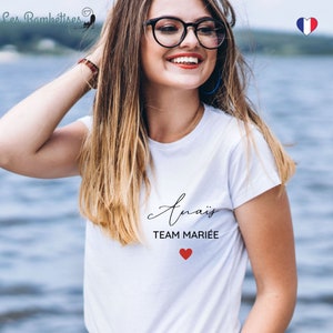 May include: White t-shirt with the text "Ana&iuml;s Team Mari&eacute;e" and a red heart. The shirt is worn by a person with long blonde hair and glasses.