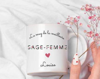 May include: White ceramic mug with the text "Le mug de la meilleure SAGE-FEMME" and the name "Louise". The word "SAGE-FEMME" is decorated with floral designs. A pink heart is below the text. The mug is on a white surface.