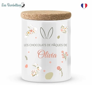 Peut inclure: Pot en céramique blanche avec un couvercle en liège. Le pot a un motif floral avec des lapins et le texte "LES CHOCOLATS DE PÂQUES DE Olivia".
