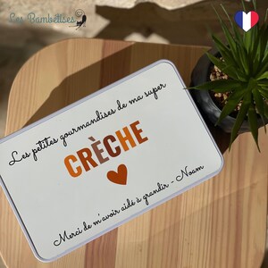May include: A white rectangular tin with the words "Les petites gourmandises de ma super CR&Egrave;CHE" in black and orange lettering. A small orange heart is below the word "CR&Egrave;CHE". The text "Merci de m'avoir aid&eacute; &agrave; grandir - Noam" is written at the bottom.