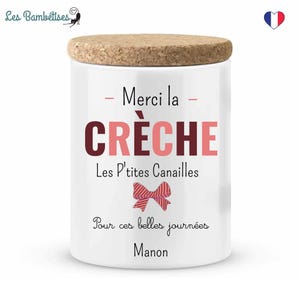 Peut inclure: Un pot en céramique blanc avec un couvercle en liège. Le pot porte l'inscription "Merci la CRÈCHE" en rose et bordeaux, ainsi que "Les P'tites Canailles" et "Pour ces belles journées Manon". Un motif de nœud rose est également présent.