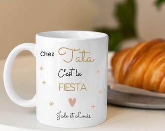 Op de afbeelding: Witte keramische mok met zwarte tekst die "Chez Tata C'est la FIESTA Jade et Louis" luidt. De tekst is in een gouden lettertype met een roze hart in het midden. De mok staat op een wit oppervlak.