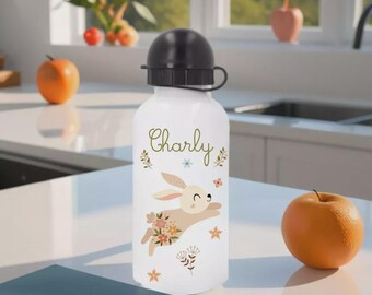 Könnte beinhalten: Weiße Wasserflasche mit schwarzem Deckel, mit dem Namen "Charly" in grüner Schrift. Ein Cartoon-Hase mit floralen Akzenten ist auf der Flasche abgebildet. Ein oranger Apfel und ein blaues Buch liegen auf der Theke.